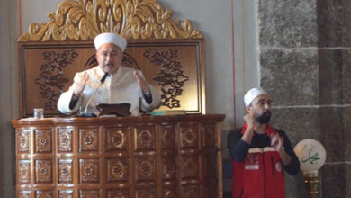 Fotoğrafta bir cami içinde yer alan, ahşap bir minberde duran bir din görevlisi görülüyor. Üzerinde beyaz cübbe ve sarık bulunan bu kişi, konuşma yaparken elleriyle bir şeyler anlatıyor. Minberin oymalı ve süslemeli bir yapısı var. Din görevlisinin yanında, kırmızı yelek giymiş ve başında beyaz bir başlık bulunan bir işaret dili tercümanı dikkat çekiyor. İşaret dili tercümanı, din görevlisinin söylediklerini işitme engellilere aktarıyor gibi görünüyor. Ortam bir caminin iç mekanına ait, dini bir etkinlik düzenleniyor.