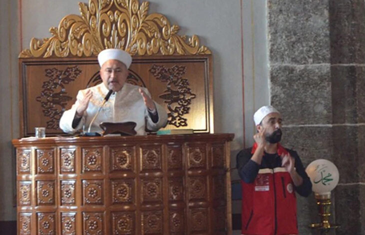 Fotoğrafta bir cami içinde yer alan, ahşap bir minberde duran bir din görevlisi görülüyor. Üzerinde beyaz cübbe ve sarık bulunan bu kişi, konuşma yaparken elleriyle bir şeyler anlatıyor. Minberin oymalı ve süslemeli bir yapısı var. Din görevlisinin yanında, kırmızı yelek giymiş ve başında beyaz bir başlık bulunan bir işaret dili tercümanı dikkat çekiyor. İşaret dili tercümanı, din görevlisinin söylediklerini işitme engellilere aktarıyor gibi görünüyor. Ortam bir caminin iç mekanına ait, dini bir etkinlik düzenleniyor.