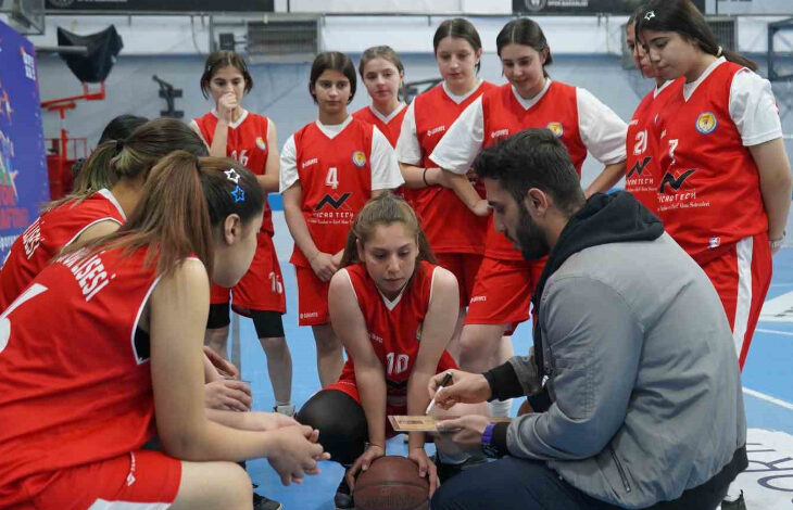 Fotoğrafta, kapalı bir spor salonunda duvar kenarına yakın bir alanda dizin üzerine çökmüş erkek antrenör ve çevresinde yaşları 13-15 arasında değişen kız sporcular var. Antrenör elindeki kağıtla yazılı taktikleri sporcularına anlatıyor.