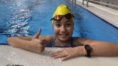 Görselde bir yüzme havuzunun kenarında duran, sarı bir bone ve yüzücü gözlüğü takmış genç bir sporcu görülüyor. Sporcu, suyun içinde, dirseklerini havuz kenarına dayamış ve sağ elini kaldırarak başparmağıyla "tamam" işareti yapıyor. Yüzünde geniş bir gülümseme var. Sağ kolunda turuncu kayışı olan bir saat takılı. Arka planda havuzun uzunluğu boyunca uzanan çizgiler ve uzak mesafede birkaç havuz kenarı detayları seçiliyor. Ortam oldukça aydınlık ve düzenli görünüyor.