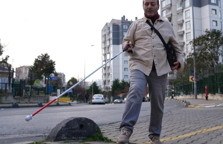 Fotoğrafta görme engelli birey Serhat Esen, elindeki beyaz baston ile kaldırımda yürümeye çalışıyor. Kaldırımın uç kısmında koruyucu amaçla yapılmış mantar şeklinde büyük bir taş var.