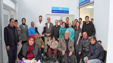 Görselde, bir sağlık merkezi veya hastane ortamında çekilmiş kalabalık bir grup fotoğrafı yer alıyor. Ön planda tekerlekli sandalyede oturan bireyler bulunurken, arka planda doktorlar, hemşireler ve diğer sağlık personeli de dahil olmak üzere çeşitli kişiler ayakta poz veriyor. Duvarlarda "Periodontoloji Lokal Müdahale" ve "Periodontoloji A.D." yazılı tabelalar görülüyor.