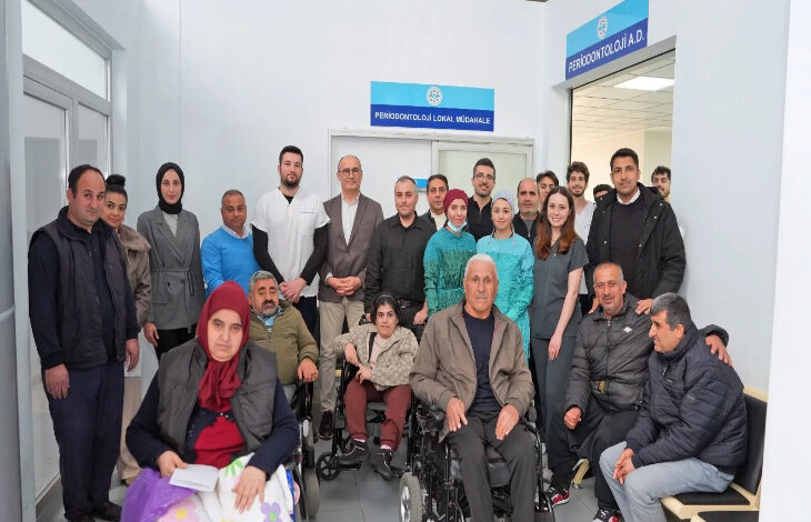 Görselde, bir sağlık merkezi veya hastane ortamında çekilmiş kalabalık bir grup fotoğrafı yer alıyor. Ön planda tekerlekli sandalyede oturan bireyler bulunurken, arka planda doktorlar, hemşireler ve diğer sağlık personeli de dahil olmak üzere çeşitli kişiler ayakta poz veriyor. Duvarlarda "Periodontoloji Lokal Müdahale" ve "Periodontoloji A.D." yazılı tabelalar görülüyor.