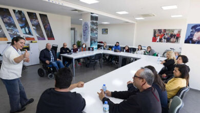 Görselde, geniş ve aydınlık bir salonda uzun U şeklinde dizilmiş masaların etrafında oturan bir grup insan görülüyor. Masalarda su şişeleri bulunuyor. Ön tarafta beyaz tişört giymiş bir kadın, el hareketleriyle bir şeyler anlatıyor. Katılımcılar dikkatle dinliyor ve bazıları el hareketleriyle karşılık veriyor. Arka planda duvarda çeşitli fotoğraflar ve afişler yer almakta. Katılımcılar arasında tekerlekli sandalye kullanan bireyler de bulunuyor.