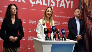 Görselde bir basın toplantısı düzenleniyor. Ortada Aylin Nazlıaka konuşma yaparken, önünde çeşitli medya kuruluşlarına ait mikrofonlar bulunuyor. Nazlıaka’nın sağında ve solunda iki kişi daha ayakta duruyor. Arka planda kırmızı bir fon üzerinde "ENGELLİLİK ÇALIŞTAYI" yazısı ve ilgili logolar yer alıyor. Atmosfer resmi ve ciddi.