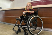 Fotoğrafta, tekerlekli sandalyede oturan bir kadın görülüyor. Sarı saçlı ve gözlüklü olan kadın, kırmızı-siyah desenli kısa kollu bir elbise giymiş. Sol kolu protez olup sağ elinde bir kağıt parçası tutuyor. Arka planda "Ahmed Adnan Saygun Sanat Merkezi" yazılı bir tabela bulunan modern bir resepsiyon alanı yer alıyor.