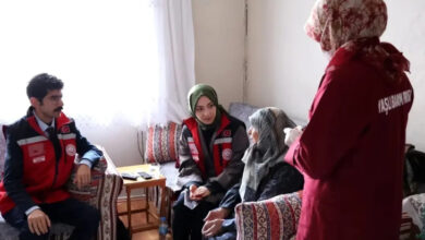 Görselde, kırmızı yelek giyen sosyal hizmet görevlileri, yaşlı bir kadını evinde ziyaret ediyor. Bir görevli sağlık hizmeti sunarken, diğerleri dikkatle dinliyor. Yaşlı kadın başörtülü ve geleneksel kıyafetler giymiş, oturma odasında bir kanepeye oturuyor.