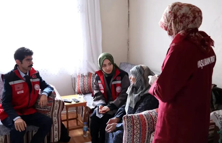 Görselde, kırmızı yelek giyen sosyal hizmet görevlileri, yaşlı bir kadını evinde ziyaret ediyor. Bir görevli sağlık hizmeti sunarken, diğerleri dikkatle dinliyor. Yaşlı kadın başörtülü ve geleneksel kıyafetler giymiş, oturma odasında bir kanepeye oturuyor.