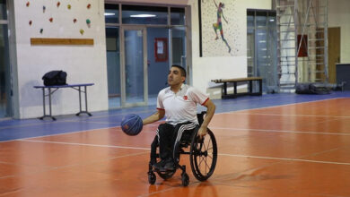 Görselde, tekerlekli sandalyede bulunan bir erkek sporcu, kapalı bir spor salonunda basketbol oynuyor. Beyaz ve kırmızı detaylı bir forma giymiş olan sporcu, elinde mavi bir basketbol topu tutuyor ve odaklanmış bir şekilde ileriye bakıyor. Zemini turuncu ve mavi olan salonun arka planında bir tırmanma duvarı, merdiven ve duvarda basketbol oynayan bir sporcunun resmi görülüyor.