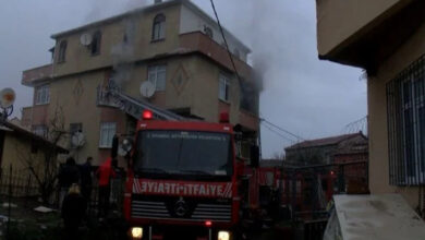 Görselde, bir apartmanın üst katından dumanlar yükseldiği görülüyor. Ön planda ise "İTFAİYE" yazılı kırmızı renkli bir itfaiye aracı bulunuyor. İtfaiye merdiveni binaya uzatılmış durumda. Çevrede birkaç kişi ve itfaiye ekipleri yangına müdahale ediyor. Hava kapalı ve ortam karanlık görünüyor.