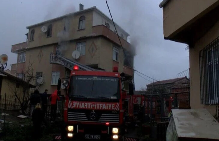 Görselde, bir apartmanın üst katından dumanlar yükseldiği görülüyor. Ön planda ise "İTFAİYE" yazılı kırmızı renkli bir itfaiye aracı bulunuyor. İtfaiye merdiveni binaya uzatılmış durumda. Çevrede birkaç kişi ve itfaiye ekipleri yangına müdahale ediyor. Hava kapalı ve ortam karanlık görünüyor.