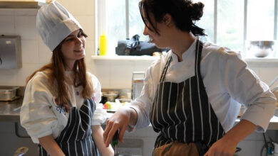 Görselde, bir mutfakta aşçılık eğitimi alan iki genç kadın görülüyor. İkisi de siyah-beyaz çizgili önlük giymiş, biri beyaz bir aşçı şapkası takıyor. Kadınlardan biri tavada bir şeyler pişirirken diğeri gülümseyerek ona bakıyor. Arka planda mutfak ekipmanları ve büyük bir pencere bulunuyor.