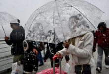 Görselde, yoğun kar yağışı altında şeffaf bir şemsiye taşıyan bir kadın görülüyor. Üzerinde beyaz bir kaban var ve yanında pembe bir valiz taşıyor. Şemsiye karla kaplanmış durumda. Arka planda, diğer yayalar da kar yağışından korunmaya çalışıyor, bazıları şemsiye kullanıyor, bazıları ise kalın kıyafetler giymiş. Ortamın bir köprü ya da sahil hattına yakın bir yer olduğu anlaşılıyor.