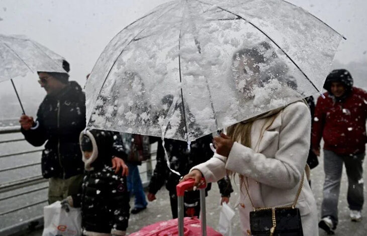 Görselde, yoğun kar yağışı altında şeffaf bir şemsiye taşıyan bir kadın görülüyor. Üzerinde beyaz bir kaban var ve yanında pembe bir valiz taşıyor. Şemsiye karla kaplanmış durumda. Arka planda, diğer yayalar da kar yağışından korunmaya çalışıyor, bazıları şemsiye kullanıyor, bazıları ise kalın kıyafetler giymiş. Ortamın bir köprü ya da sahil hattına yakın bir yer olduğu anlaşılıyor.