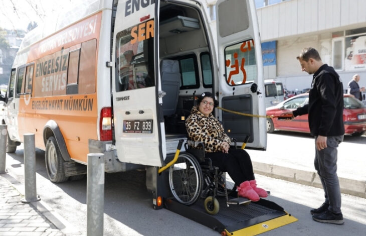 Fotoğrafta, tekerlekli sandalyedeki bir kadın Konak Belediyesi’nin "Engelsiz Servis" aracına biniyor. Araç, engelli bireyler için özel olarak tasarlanmış bir rampaya sahip. Kadın, leopar desenli montu ve pembe terlikleriyle mutlu bir şekilde kameraya bakıyor. Yanında ise ona yardımcı olan bir görevli bulunuyor.