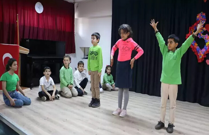 Görselde, bir grup ilkokul öğrencisi sahnede tiyatro oyunu sergiliyor. Çocuklardan bazıları ayakta rollerini canlandırırken, diğerleri sahnede yere oturmuş halde oyunu takip ediyor. Sahnenin arka planında kırmızı perde, piyano ve bir Türk bayrağı yer alıyor. Öğrencilerin kıyafetleri genellikle yeşil ve beyaz tonlarında, ancak pembe giyen bir öğrenci dikkat çekiyor.