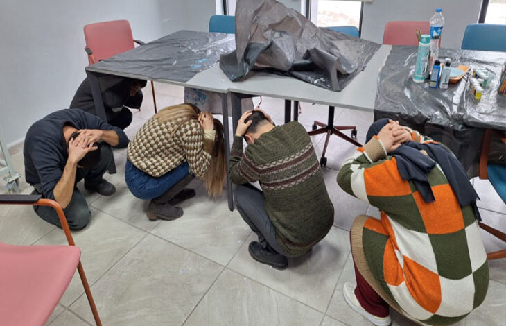 Fotoğrafta, beş kişi bir odada deprem tatbikatı yapıyor. Katılımcılar diz çökmüş, başlarını elleriyle koruyarak siper almış durumda. Ortada bir masa var ve üstü plastik örtüyle kaplanmış. Çevrede farklı renkte sandalyeler ve masanın üzerinde su şişesi, temizlik malzemeleri gibi çeşitli eşyalar bulunuyor. Oda geniş ve aydınlık, zemini seramik kaplı. Tatbikat, deprem anında güvenli pozisyon alma eğitimini gösteriyor.