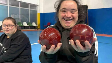 Görselde bir spor salonunda iki kişi görülüyor. Ön planda, siyah eşofman giyen bir kişi elinde iki kırmızı bocce topu tutuyor ve gülümseyerek poz veriyor. Arka planda başka bir kişi daha oturuyor. Salonun zemininde mavi renkli spor matları bulunuyor.