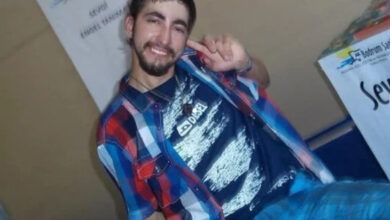 Görselde, genç bir adam kameraya gülümseyerek poz veriyor. Üzerinde kareli bir gömlek ve desenli bir tişört bulunuyor.