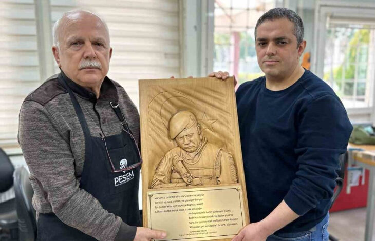 Fotoğrafta, iki adam bir ahşap kabartma portreyi birlikte tutuyor. Soldaki kişi önlüğüyle ahşap işçiliğiyle ilgilendiğini gösterirken, sağdaki kişi ise sade bir kıyafet giymiş. Ahşap eser, şehit Ömer Halisdemir’in kabartma portresi ve alt kısmında bir yazı içeriyor. Arka planda atölye ortamına benzeyen bir mekân bulunuyor.