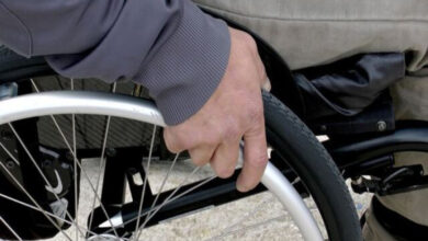 Görselde, bir kişinin tekerlekli sandalyesinin tekerleğini eliyle ittiği görülüyor. Kişi koyu renkli bir ceket ve açık renkli bir pantolon giymiş. Arka planda ise bulanık bir zemin bulunuyor.