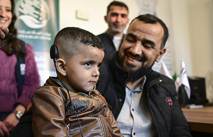 Fotoğrafta, işitme cihazı takılı küçük bir çocuk ve ona gülümseyerek bakan bir adam görülüyor. Çocuk kahverengi bir ceket giyerken, adam siyah mont ve açık renkli bir gömlek giymiş. Arkalarında birkaç kişi daha bulunuyor ve ortam resmi bir kurum ya da sağlık merkezi gibi görünüyor. Çocuğun ciddi bir ifadesi varken, yanındaki adam mutlu ve gururlu bir şekilde gülümsüyor.