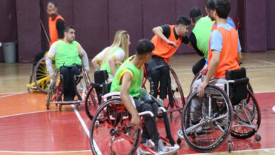 Bu görselde, Astor Enerji Şanlıurfa Bedensel Engelliler Spor Kulübü oyuncuları antrenman yapıyor. Sporcular, tekerlekli sandalyeleriyle sahada hareket halinde ve oyuna odaklanmış görünüyorlar. Üzerlerindeki yeşil ve turuncu yelekler, antrenman sırasında takımlara ayrıldıklarını gösteriyor.