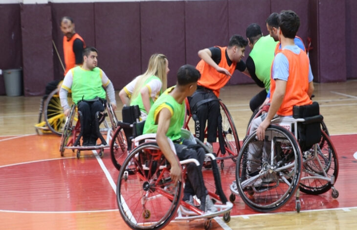 Bu görselde, Astor Enerji Şanlıurfa Bedensel Engelliler Spor Kulübü oyuncuları antrenman yapıyor. Sporcular, tekerlekli sandalyeleriyle sahada hareket halinde ve oyuna odaklanmış görünüyorlar. Üzerlerindeki yeşil ve turuncu yelekler, antrenman sırasında takımlara ayrıldıklarını gösteriyor.