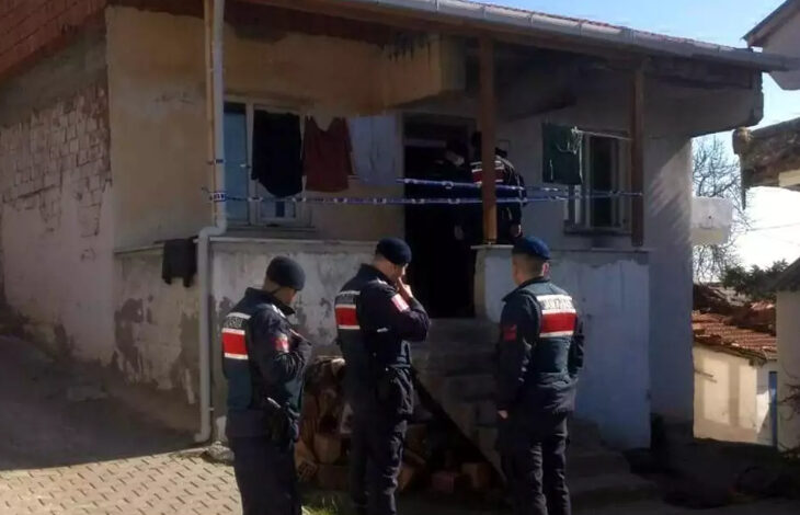 Görselde, tek katlı eski bir evin önünde görev yapan jandarma ekipleri görülüyor. Üç jandarma personeli siyah bere ve üniformalarıyla dikkat çekerken, biri telsiziyle iletişim kuruyor. Evin giriş kapısına mavi-beyaz şerit çekilmiş, içeride ise birkaç görevli daha bulunuyor. Ev eski ve yıpranmış görünüyor, duvarlarında çatlaklar var. Merdivenlerin altında odun yığınları yer alıyor.