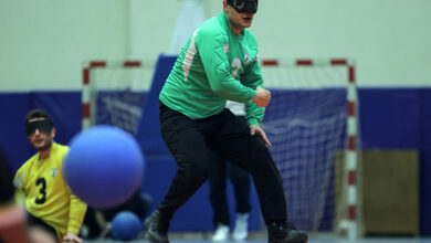 Görselde, bir goalball oyuncusu oyun esnasında hareket halinde görülüyor. Yeşil forma giyen sporcu, siyah göz bandı takıyor ve yere yaklaşarak mavi goalball topunu takip ediyor. Arka planda bir başka oyuncu, sarı forma içinde ve göz bandıyla görülüyor. Salon sporlarına uygun bir zeminde, kale filesi ve mavi minderlerle çevrili bir oyun alanı bulunuyor.