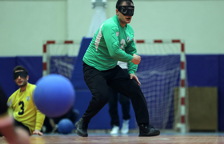 Görselde, bir goalball oyuncusu oyun esnasında hareket halinde görülüyor. Yeşil forma giyen sporcu, siyah göz bandı takıyor ve yere yaklaşarak mavi goalball topunu takip ediyor. Arka planda bir başka oyuncu, sarı forma içinde ve göz bandıyla görülüyor. Salon sporlarına uygun bir zeminde, kale filesi ve mavi minderlerle çevrili bir oyun alanı bulunuyor.