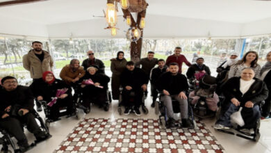 Görselde, bir grup insan kapalı ve aydınlık bir mekânda bir araya gelmiş. Çoğunluğu tekerlekli sandalyede bulunan bireyler, mutlu bir şekilde poz veriyor. Ortada ahşap detaylı bir sütun ve asılı lambalar bulunuyor. Grup, sıcak bir atmosferde, dostane ve samimi bir etkinlikte bir araya gelmiş gibi görünüyor.