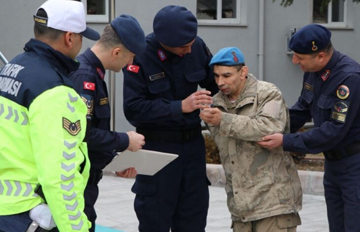 Fotoğrafta, jandarma ve trafik polisleri üniformalı bir grup görevli, asker kıyafeti giymiş bir engelli bireye yardımcı oluyor. Engelli birey mavi bereli ve kamuflajlı, yanında duran jandarmalar ona destek vererek eline bir belge veya madalya tutuşturuyor. Görevlilerden biri elinde bir dosya tutuyor. Arka planda gri bir bina görülüyor. Fotoğraf, engelli bireyin askerlik hayalinin gerçekleştiği özel bir anı yansıtıyor.