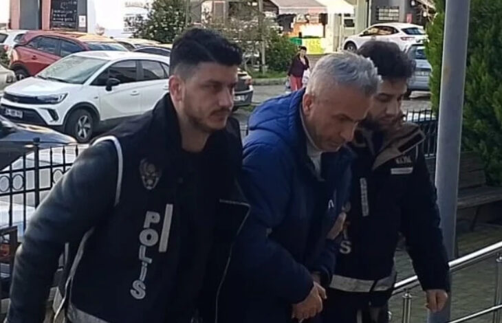 Görselde, polis ekipleri kelepçeli bir adamı kolluk kuvvetleri eşliğinde adliyeye veya karakola götürüyor. Adam koyu renkli bir mont giymiş ve başı öne eğik durumda. Polis memurları "POLİS" yazılı yelekler giymiş ve güvenlik önlemleri almış. Arka planda ise araçlar ve şehir ortamı görülüyor.