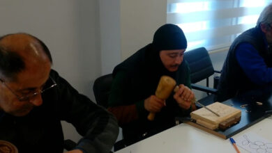 Fotoğrafta bir atölye çalışması görülüyor. Üç kişi, masa üzerinde bulunan ahşap parçalar üzerine oyma işlemi yapıyor. Ortadaki kişi siyah başörtü ve koyu renkli kıyafet giyerken, elinde bir keski ve tokmak tutarak ahşap üzerinde çalışıyor. Soldaki kişi gözlüklü ve odaklanmış bir şekilde çalışırken, sağdaki kişi ise mavi yelek giymiş ve ahşap oymacılığı ile ilgileniyor.