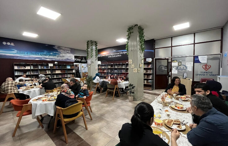 Görselde, bir kütüphane veya okuma salonu ortamında düzenlenen bir iftar yemeği görülüyor. Masalarda iftar için çeşitli yemekler yer alırken, farklı yaş gruplarından insanlar bir arada yemeklerini yiyor ve sohbet ediyor. Arkada kitap rafları, sütunlarda süs bitkileri ve duvarlarda anlamlı yazılar bulunuyor. Sağ tarafta "Türkiye Diyanet Vakfı" logosunun yer aldığı bir pano görülüyor.