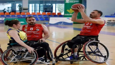 Görselde, tekerlekli sandalye basketbol oyuncuları bir spor salonunda antrenman yaparken görülüyor. Kırmızı formalar giyen iki erkek sporcu ve sarı-lacivert formaya sahip bir kadın sporcu sahada yer alıyor. Ön planda, kırmızı formalı bir oyuncu şut atmaya hazırlanırken, diğer iki sporcu dikkatle onu izliyor. Spor salonunun arka planında tribünler ve çeşitli sponsorlara ait afişler bulunuyor.