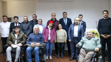 Fotoğrafta bir grup insan, kapalı bir mekânda toplu halde poz veriyor. Ön sırada tekerlekli sandalyede oturan üç kişi ve ayakta duran engelli bireyler bulunuyor. Arkalarında ise takım elbiseli ve günlük kıyafetli birkaç kişi daha yer alıyor. Sağ tarafta "T.C." ibaresinin yer aldığı bir kürsü ve mikrofon görülüyor.