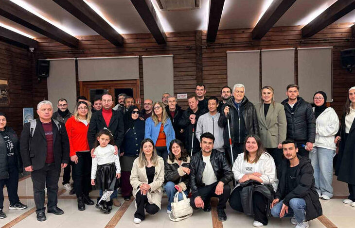 Görselde, geniş bir kapalı mekânda büyük bir grup insan birlikte poz vermektedir. Çeşitli yaş gruplarından bireyler bulunmakta ve bazıları baston kullanmaktadır. Arka planda ahşap duvarlar, tavan kirişleri ve kapalı pencereler görülmektedir. İnsanlar sıcak bir ortamda bir araya gelmiş olup, etkinlik iftar organizasyonu gibi sosyal bir buluşmaya işaret etmektedir.