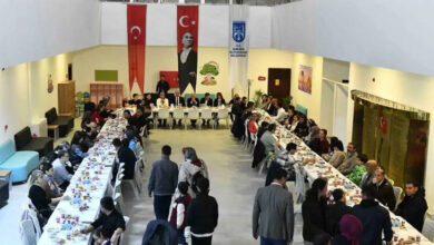 Görselde, büyük bir kapalı alanda uzun sofralar kurulmuş ve birçok kişi iftar yemeği için toplanmış. Masalarda yiyecek ve içecekler bulunuyor. Arka planda Türk bayrakları, Atatürk posteri ve Ankara Büyükşehir Belediyesi'ne ait bir afiş asılı. İnsanlar yemeklerini yerken, bazıları sohbet ediyor.