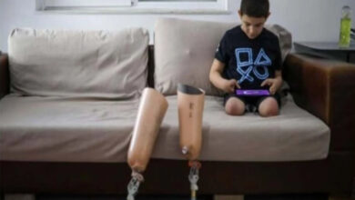 Görselde, bir çocuk evin içinde bir kanepede oturuyor ve tabletle oyun oynuyor. Üzerinde PlayStation sembollerinin bulunduğu siyah bir tişört var. Çocuğun protez bacakları kanepe önünde duruyor. Arka planda pencere, bir masa ve üzerinde bir şişe su bulunan sehpa yer alıyor.