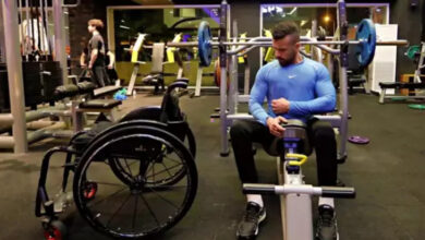 Görselde, spor salonunda antrenman yapan engelli bir birey görülüyor. Mavi uzun kollu spor tişörtü ve siyah pantolon giyen adam, ağırlık kaldırma makinesinde oturuyor ve kaslarını kontrol ediyor. Yanında tekerlekli sandalyesi bulunuyor. Arkada spor salonunun ekipmanları ve başka spor yapan kişiler görülebiliyor. Salon modern ve iyi aydınlatılmış bir ortamda bulunuyor.