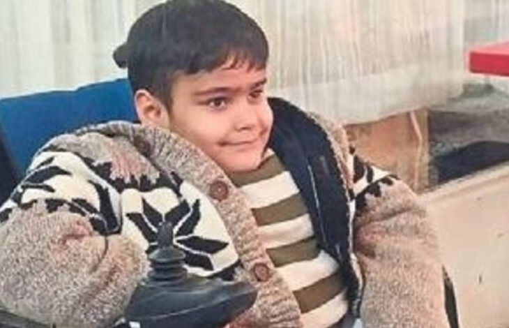 Fotoğrafta, genç bir çocuk akülü tekerlekli sandalyesinde oturuyor. Üzerinde beyaz, siyah desenli kalın bir hırka ve altına yeşil-beyaz çizgili bir kazak giymiş. Hafifçe gülümseyerek kameraya bakıyor. Arka planda perde ve pencere bulunan bir kapalı mekân yer alıyor.