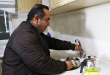 Görselde, orta yaşlı bir adam mutfakta su dolu bir çaydanlığı musluktan dolduruyor. Üzerinde kahverengi kalın bir mont bulunuyor. Tezgâhta bir demlik ve bir bez yer alıyor. Mutfak temiz ve düzenli görünüyor, üst dolaplar açık renk ahşap kaplamaya sahip.