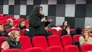 Görselde, kırmızı koltuklu bir konferans veya söyleşi salonunda gerçekleştirilen bir etkinlik yer almaktadır. Başörtülü, siyah montlu bir kadın elinde mikrofonla konuşurken, çevresindeki katılımcılar onu dinliyor. Katılımcılar arasında başörtülü ve başı açık kadınlar ile erkekler bulunuyor. Arka planda siyah ve gri karelerden oluşan bir duvar tasarımı göze çarpıyor.