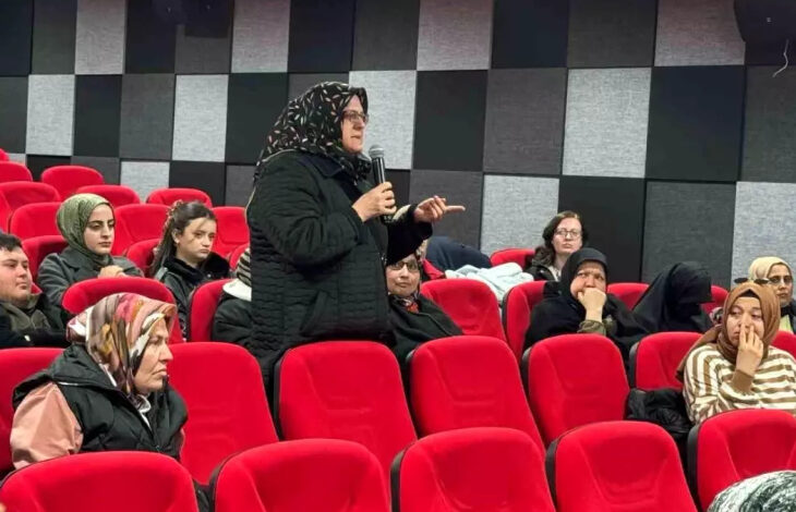 Görselde, kırmızı koltuklu bir konferans veya söyleşi salonunda gerçekleştirilen bir etkinlik yer almaktadır. Başörtülü, siyah montlu bir kadın elinde mikrofonla konuşurken, çevresindeki katılımcılar onu dinliyor. Katılımcılar arasında başörtülü ve başı açık kadınlar ile erkekler bulunuyor. Arka planda siyah ve gri karelerden oluşan bir duvar tasarımı göze çarpıyor.