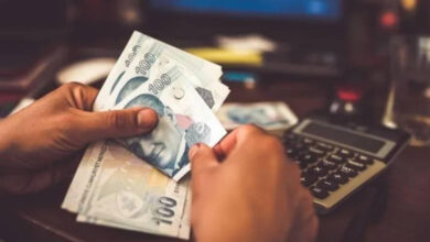 Görselde, bir kişinin elinde 100 TL’lik banknotlar bulunmaktadır. Arka planda bir hesap makinesi ve bulanık bir bilgisayar ekranı görülmektedir. Ortam, bir çalışma masası veya finansal işlemlerin yapıldığı bir alan izlenimi vermektedir.