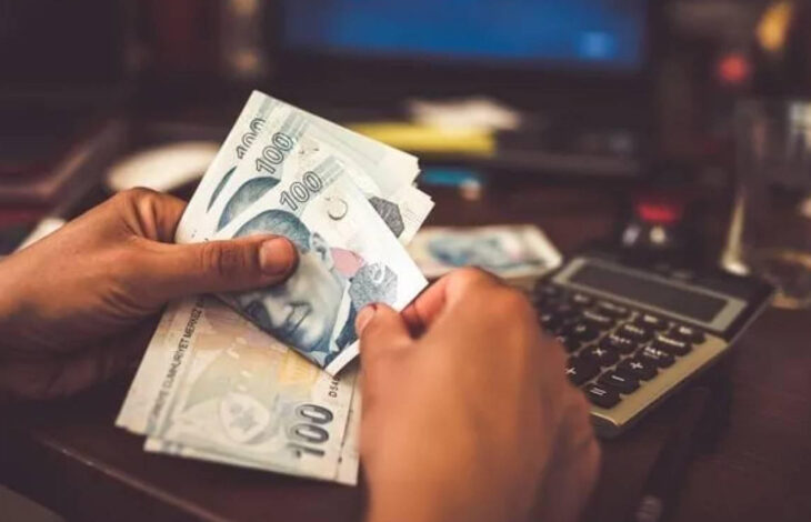 Görselde, bir kişinin elinde 100 TL’lik banknotlar bulunmaktadır. Arka planda bir hesap makinesi ve bulanık bir bilgisayar ekranı görülmektedir. Ortam, bir çalışma masası veya finansal işlemlerin yapıldığı bir alan izlenimi vermektedir.