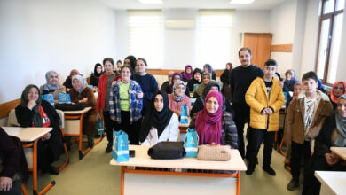 Görselde, bir sınıf ortamında toplanmış farklı yaş gruplarından kadınlar, çocuklar ve bir erkek yer alıyor. Katılımcıların bir kısmı başörtülü, bazıları ise başı açık. Ön sıralarda oturan kadınların masalarında küçük mavi hediye çantaları bulunuyor. Arka planda ise ayakta duran kişiler gülümsüyor ve kameraya bakıyor.