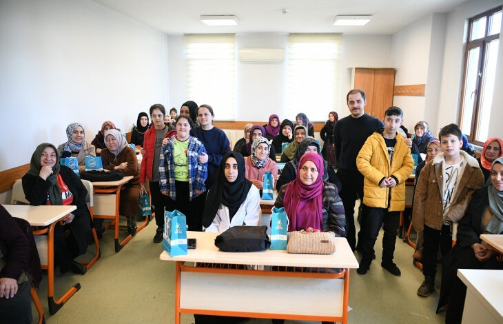 Görselde, bir sınıf ortamında toplanmış farklı yaş gruplarından kadınlar, çocuklar ve bir erkek yer alıyor. Katılımcıların bir kısmı başörtülü, bazıları ise başı açık. Ön sıralarda oturan kadınların masalarında küçük mavi hediye çantaları bulunuyor. Arka planda ise ayakta duran kişiler gülümsüyor ve kameraya bakıyor.